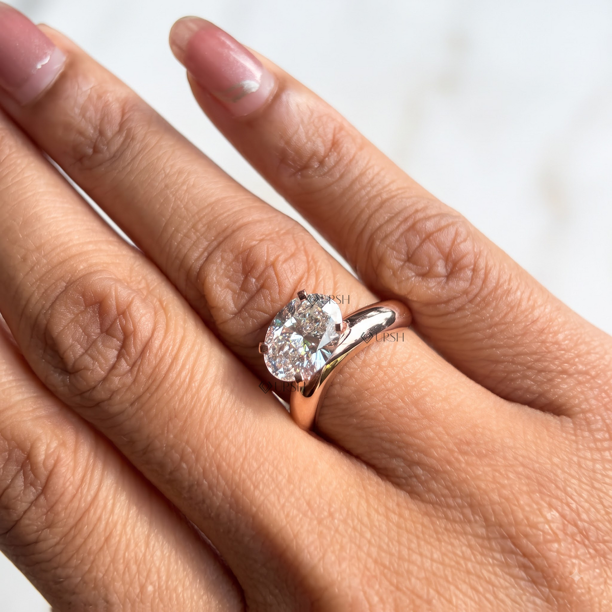 A close-up of a hand wearing a rose gold ring with a large round brilliant-cut diamond. The ring features a split shank design and is shown on a finger with pink manicured nails against a soft, out-of-focus light background.