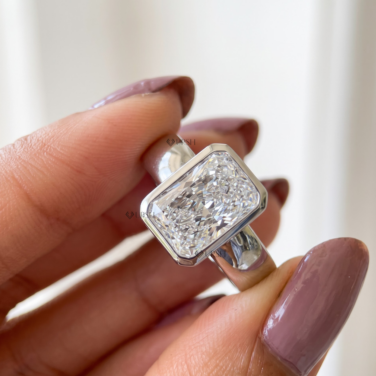 Close up of radiant cut diamond white gold bezel ring held between fingers
