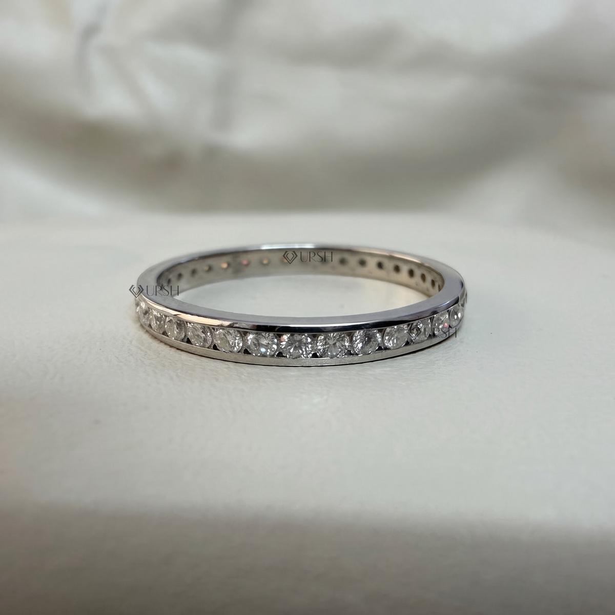 A silver eternity band ring set with small clear round-cut gemstones, resting on a soft white fabric background.