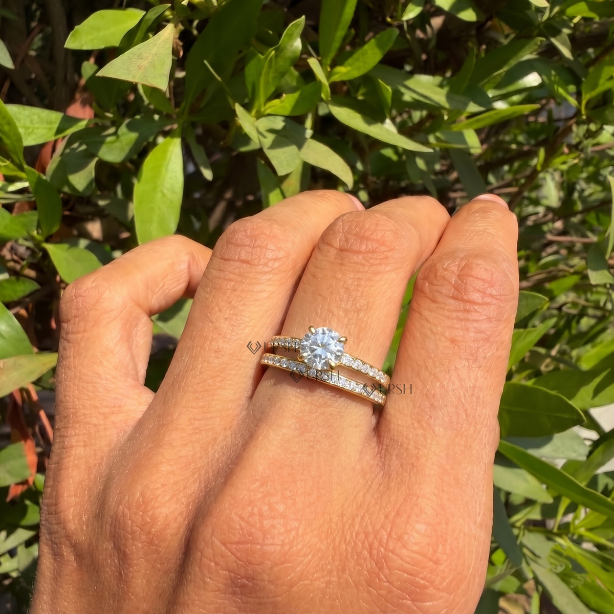 A close-up of a hand wearing a silver-toned engagement ring with a round brilliant-cut center stone and a matching pavé band, set against a background of green foliage.