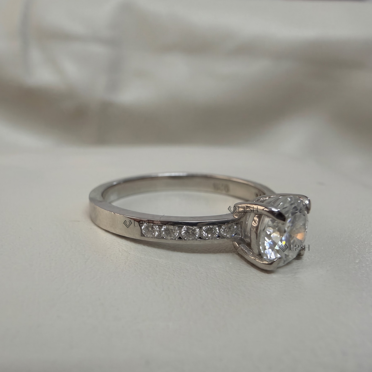 A silver-toned diamond ring with a large cushion-cut center stone and a pavé band, photographed on a soft white fabric background.