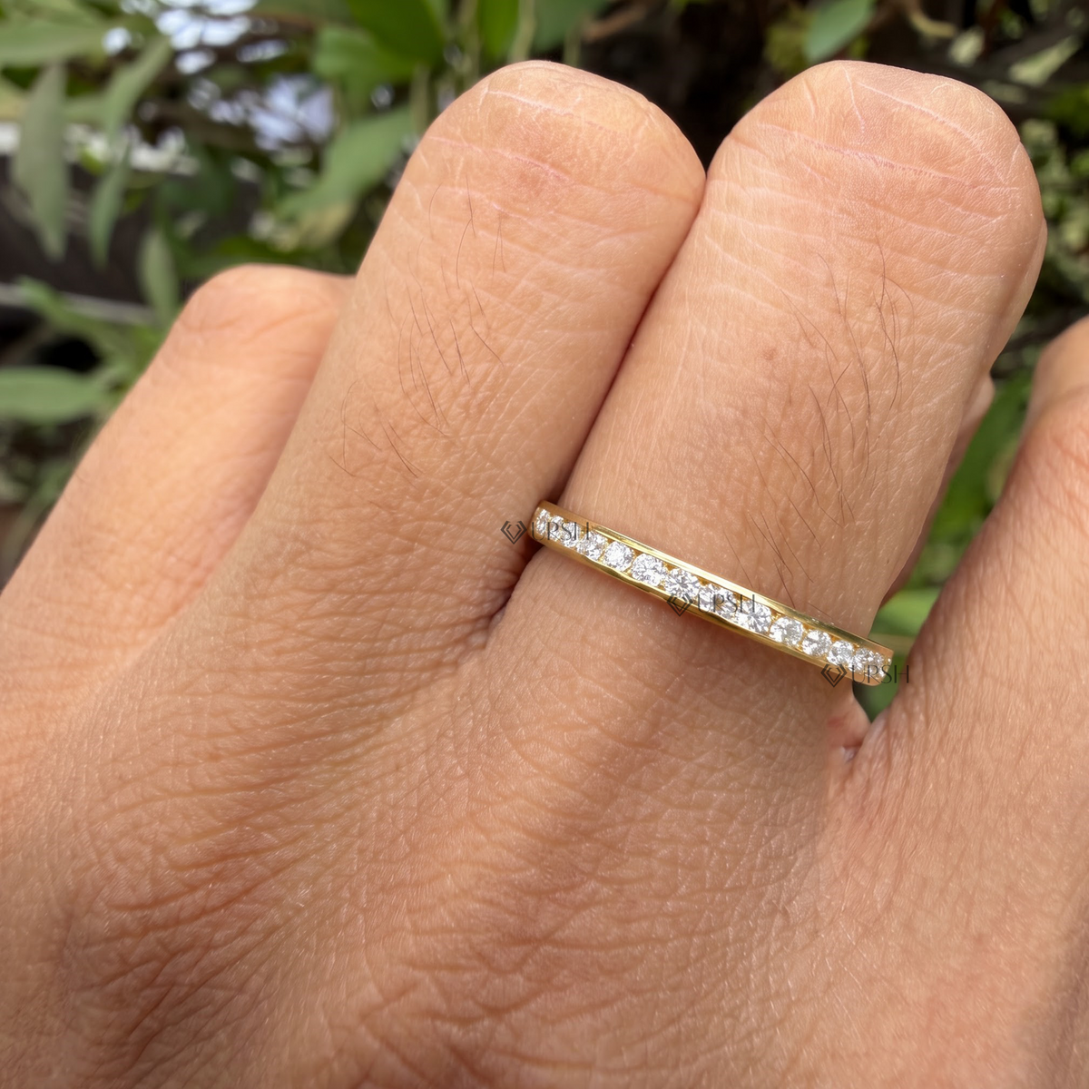 A close-up of a hand wearing a thin yellow gold eternity ring set with small clear stones on one finger, with blurred green foliage in the background.