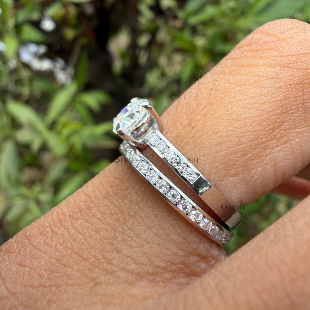A close-up of a silver engagement ring with a round-cut center stone and a matching pave-set band, worn on a person's finger with a blurred green outdoor background.
