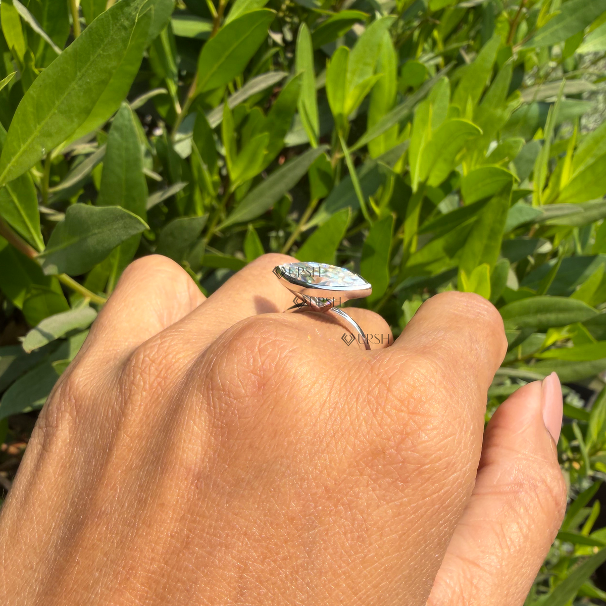 Wide daylight perspective of east-west marquise solitaire ring worn against natural garden foliage