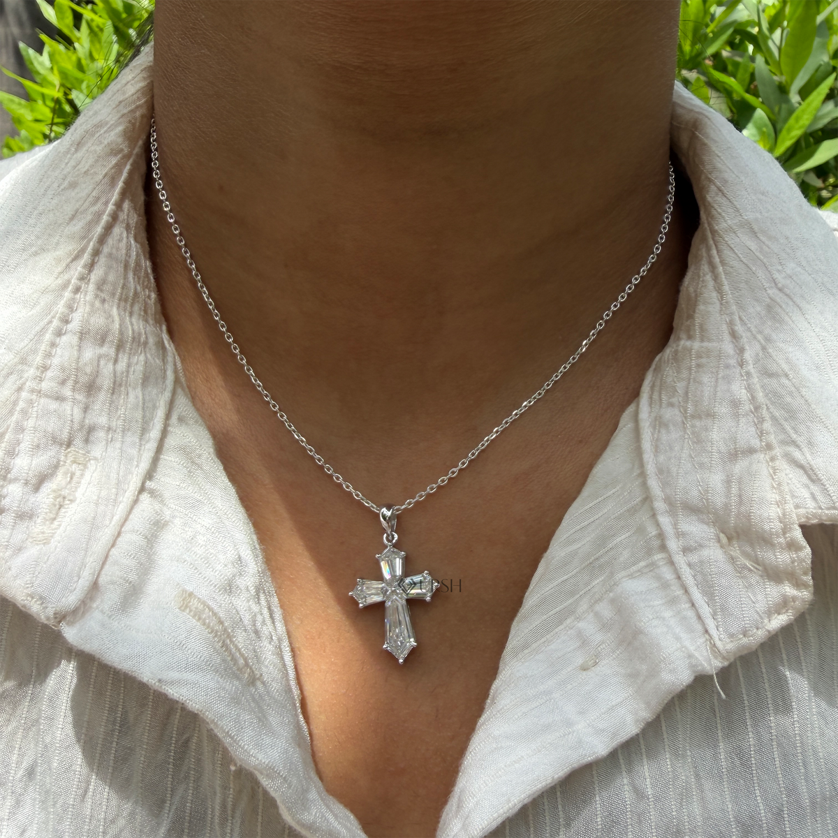 A silver cross pendant necklace worn on a person's neck, styled with a white textured linen shirt, with green foliage in the background.