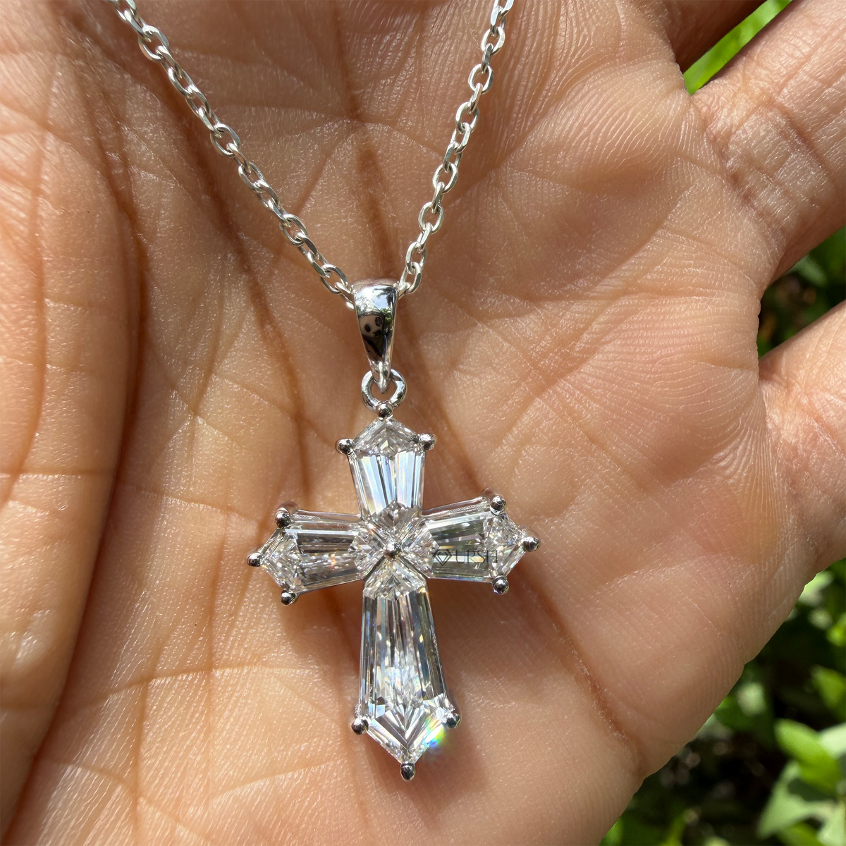A silver cross pendant with clear baguette-cut gemstones, held in a person's hand against a blurred green outdoor background.